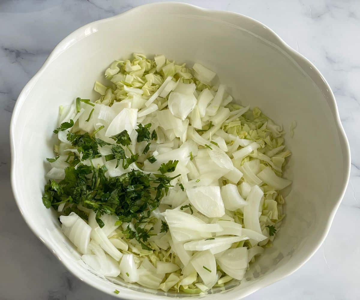 A ceramic bowl is with cabbage strips and sliced onions and chopped cilantro on the table.