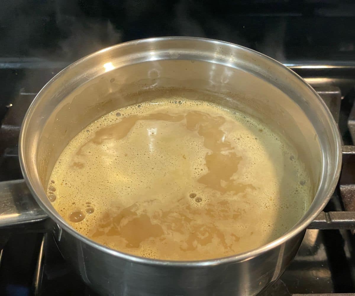 A stainless steel pot filled with light brown kashayam (herbal decoction) boiling on a gas stove, with foam and bubbles forming on the surface of the simmering liquid.