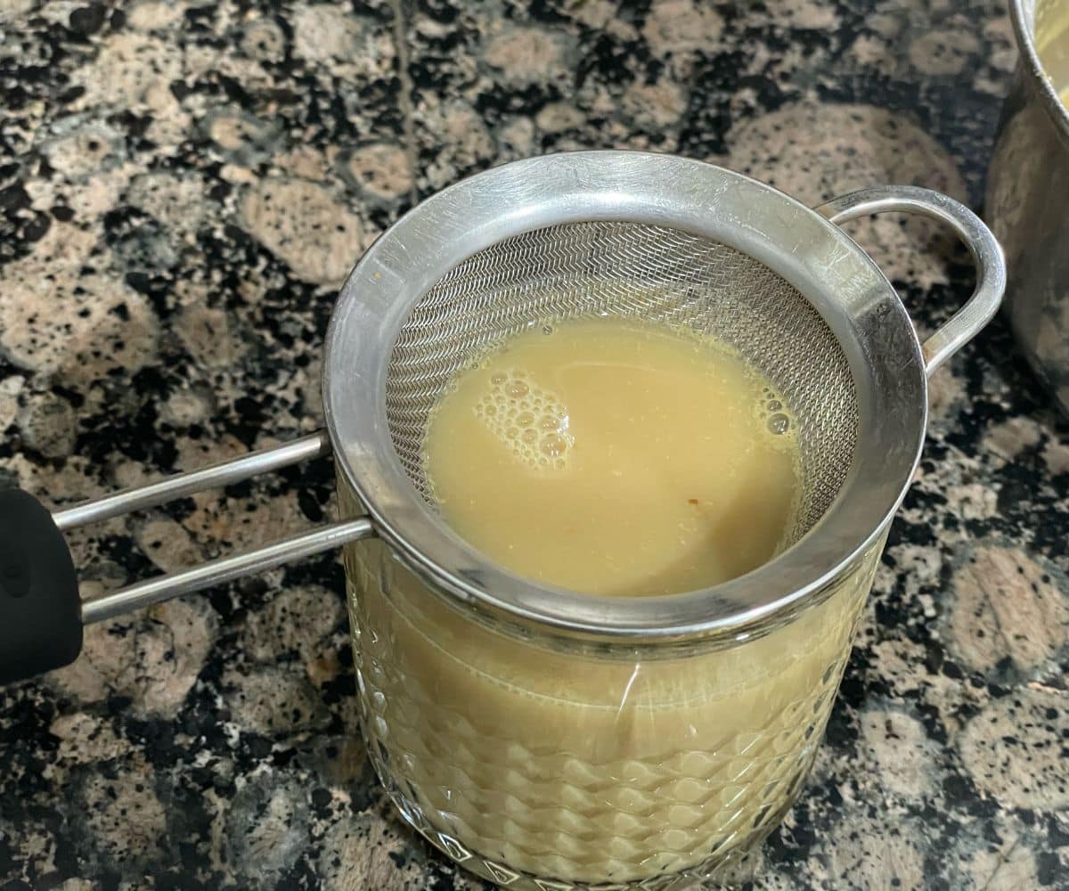 A fine mesh strainer resting over a textured glass tumbler on a granite countertop, with warm beige kashayam being strained into the glass, forming small bubbles on the surface.
