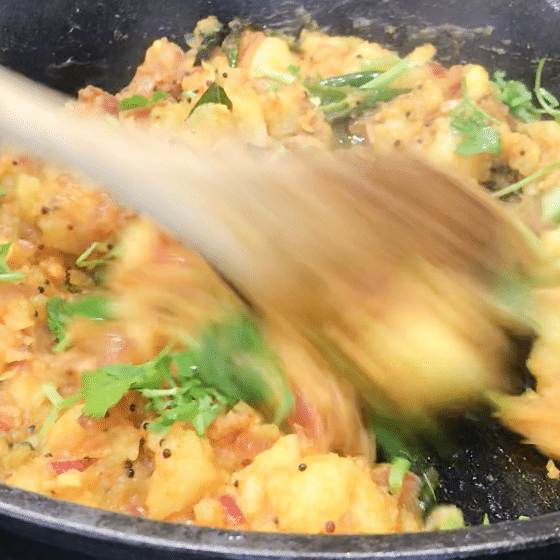 A pan filled with potato curry and a lady is stirring with a wooden spoon.