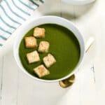 A white bowl of spinach soup is topped with croutons