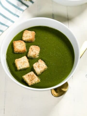 A white bowl of spinach soup is topped with croutons