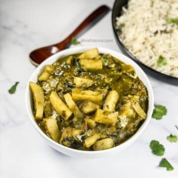 A bowl of aloo palak Curry is on the marble table with a wooden spoon by the side.