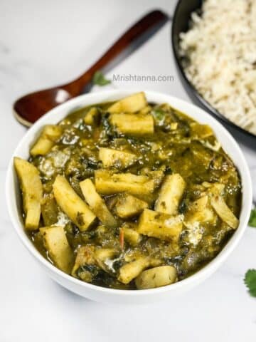 A bowl of aloo palak Curry is on the marble table with a wooden spoon by the side.
