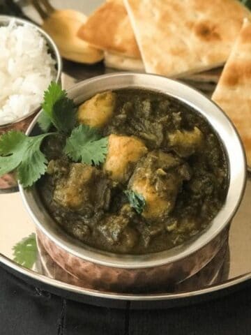 A copper bowl is filled with aloo palak curry on the steel plate.