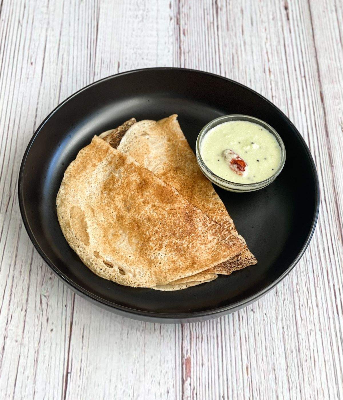 A black plate is with wheat dosa and bowl of chutney.