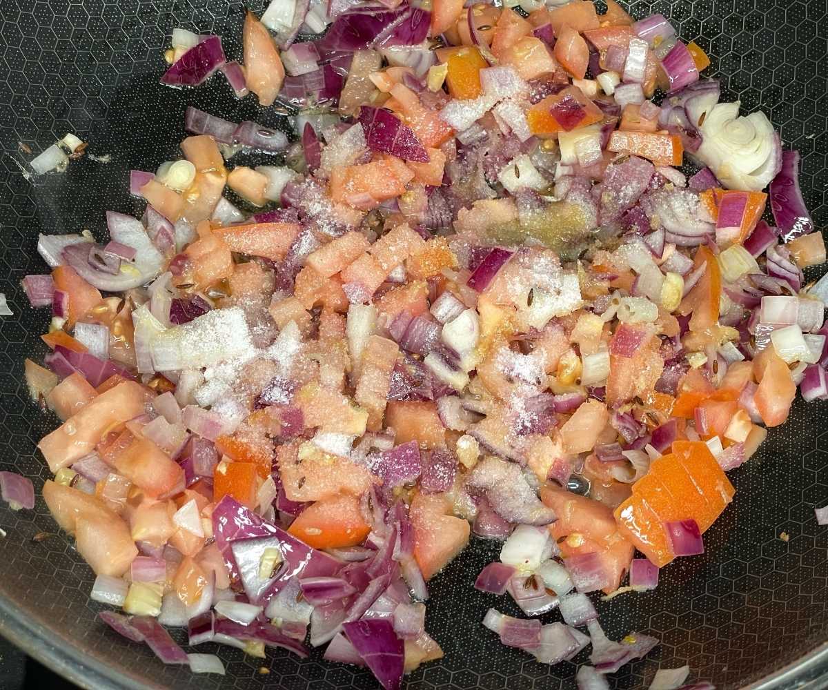 A close-up of a frying pan filled with chopped red onions, tomatoes, garlic, and spices sizzling together as the base for a curry.