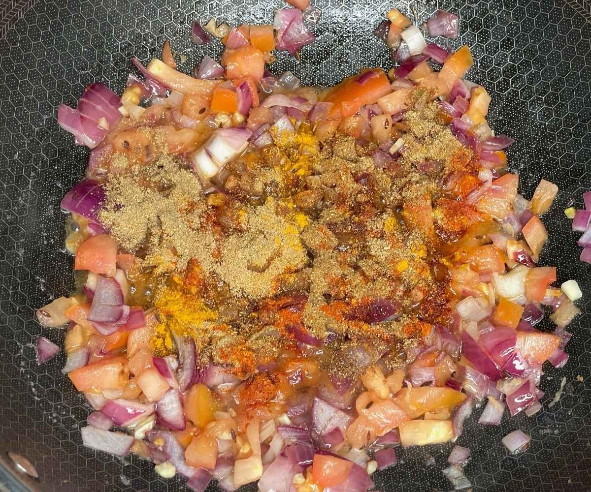 A close-up of a frying pan filled with chopped red onions, tomatoes, garlic, and spices sizzling together as the base for a curry.
