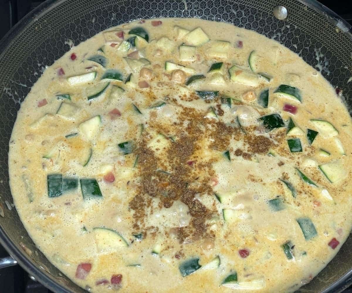 A large skillet filled with creamy zucchini curry simmers on the stove, with chunks of zucchini and spices visible on the surface.