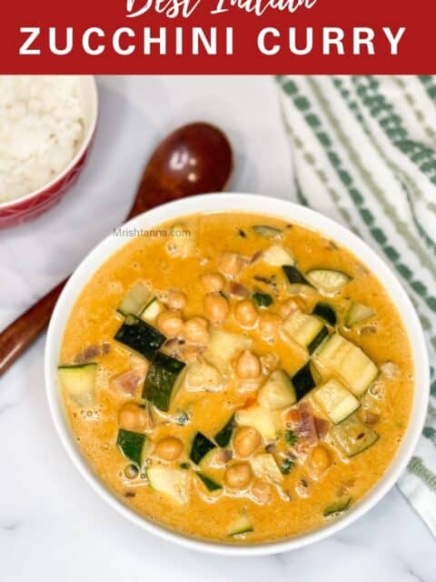 A white bowl of creamy zucchini and chickpea curry sits on a marble surface next to a red bowl of rice, a wooden spoon, and a green-and-white striped cloth.