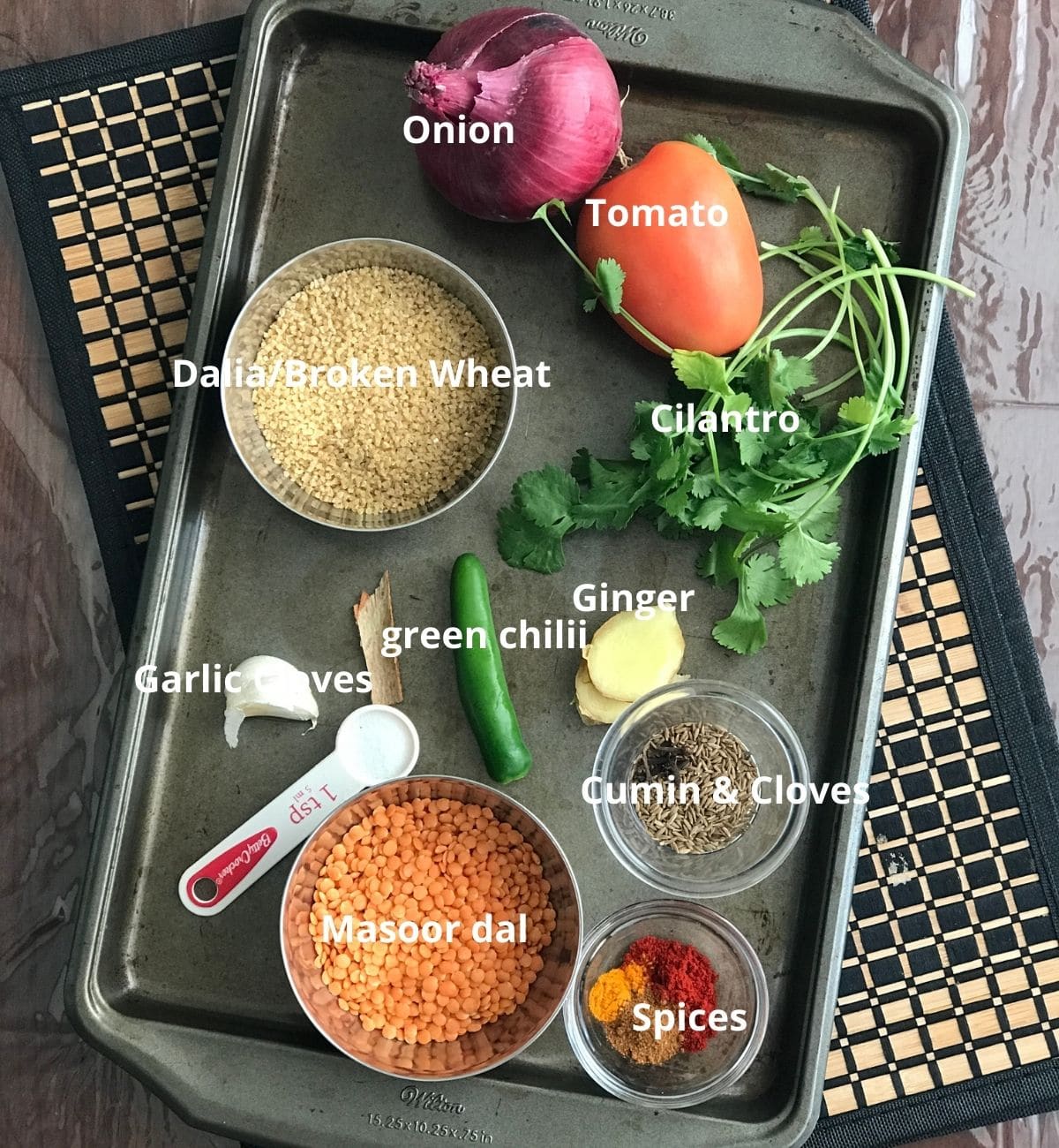 A tray with khcidi ingredients like broken wheat, masoor dal, tomato and onion