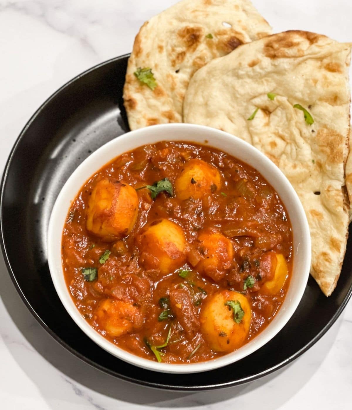 A bowl is with dum aloo and plate of naan is on the table.