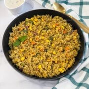 A plate of quinoa pulao is on the table with a bowl of raita and a spoon.