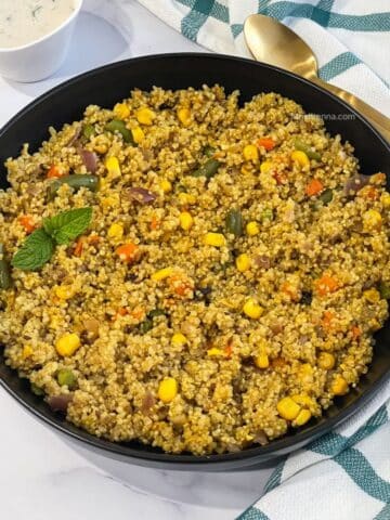 A plate of quinoa pulao is on the table with a bowl of raita and a spoon.