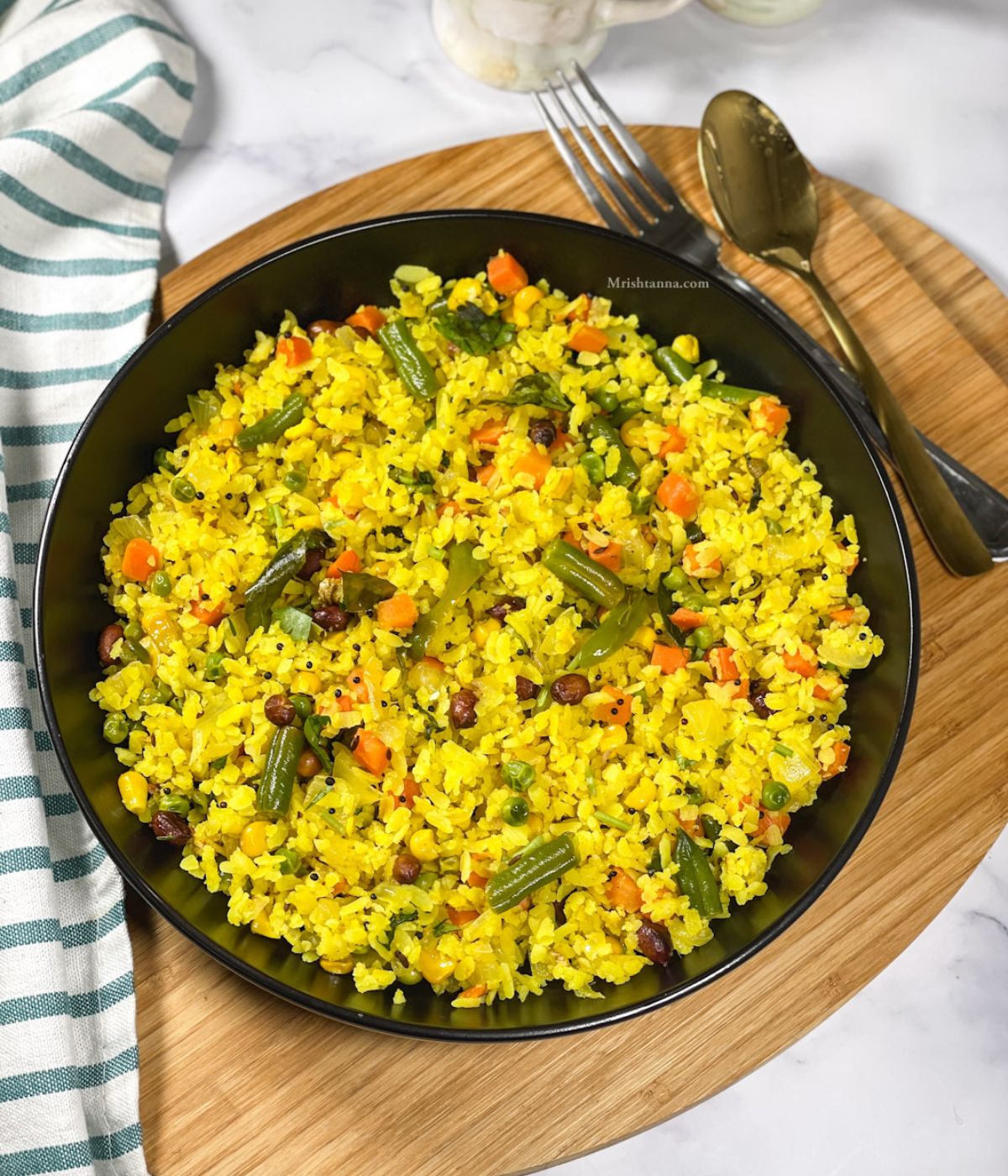 A plate of vegetable poha is on the wooden tray with a fork and spoon by the side.