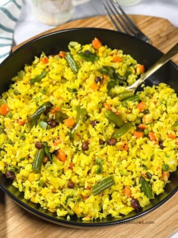 A plate of vegetable poha is on the wooden board and a spoon is inserted.