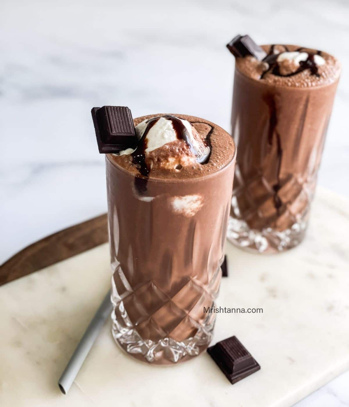Two glasses are filled with vegan chocolate milkshake and topped with ice cream and chocolate.