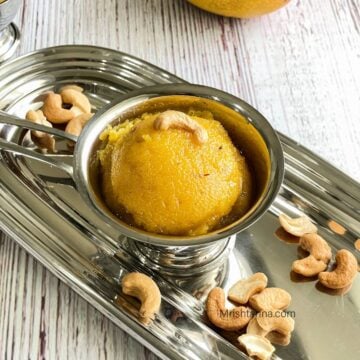 A steel cup of mango sheera is on the steel tray with spoons.