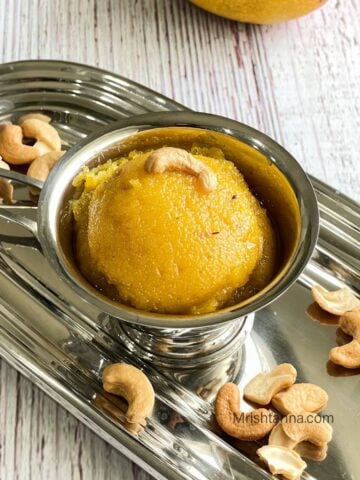 A steel cup of mango sheera is on the steel tray with spoons.