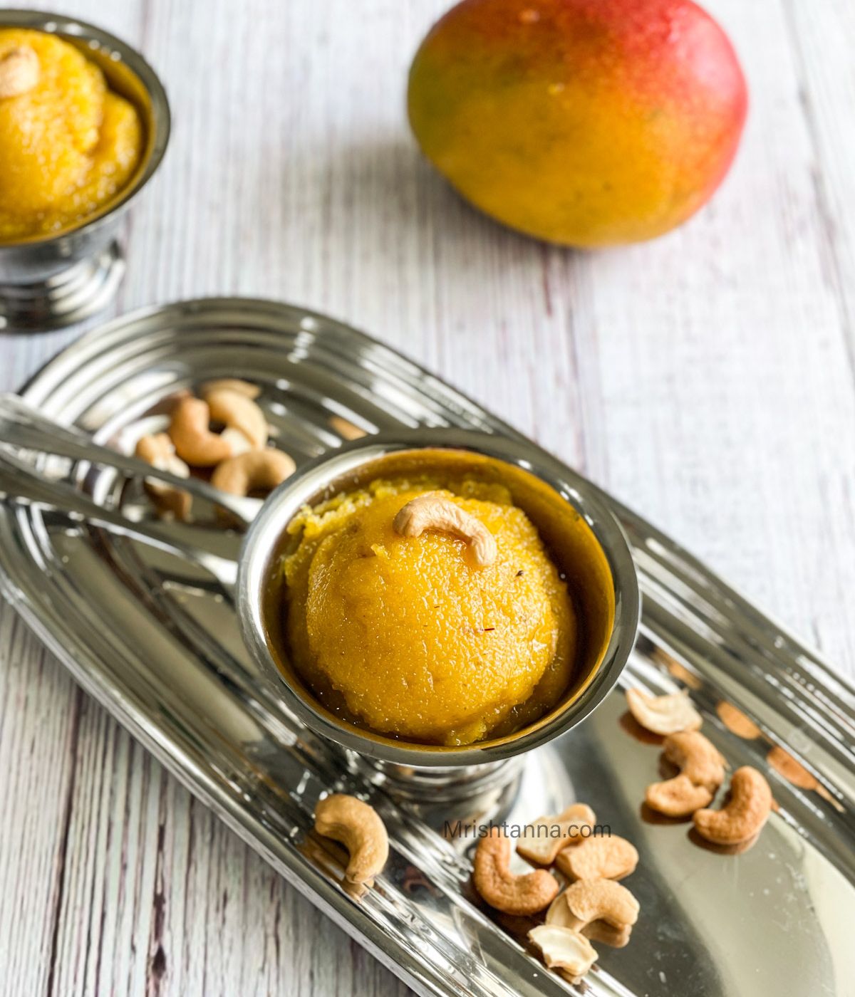 A steel cup is with mango kesari and topped with roasted cashews.