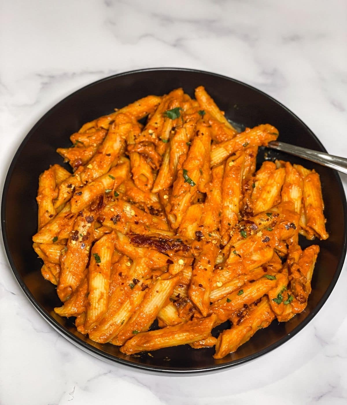 A black plate is with sun dried tomato pasta and a fork.