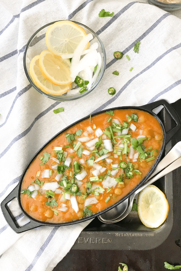 A cast iron bowl is with matar chaat and topped with onions and cilantro.