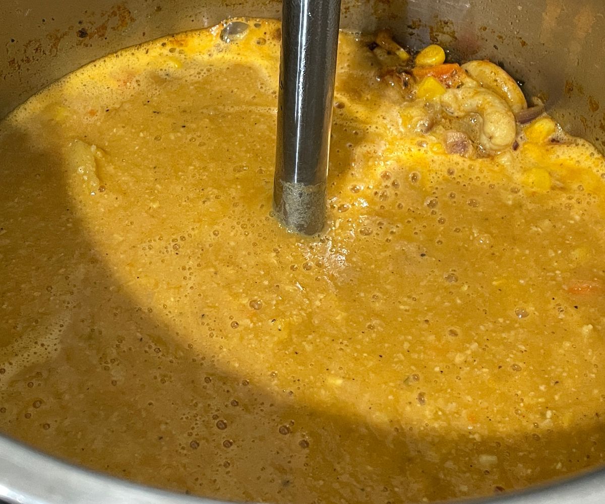 An instant pot has corn chowder and blending with an immersion blender.