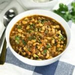 A white bowl is with black eyed peas curry along with spoons on the table