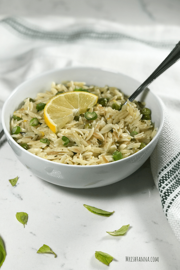 A white bowl filled with orzo and fork inserted and a lemon wedge for garnish