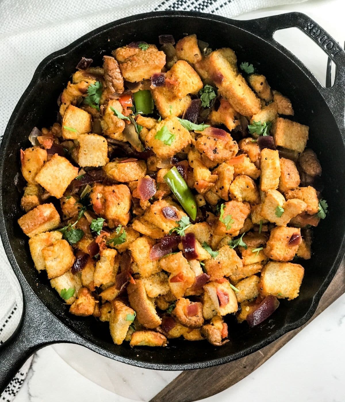 A cast iron pan is filled with bread upma and placed over the white surface