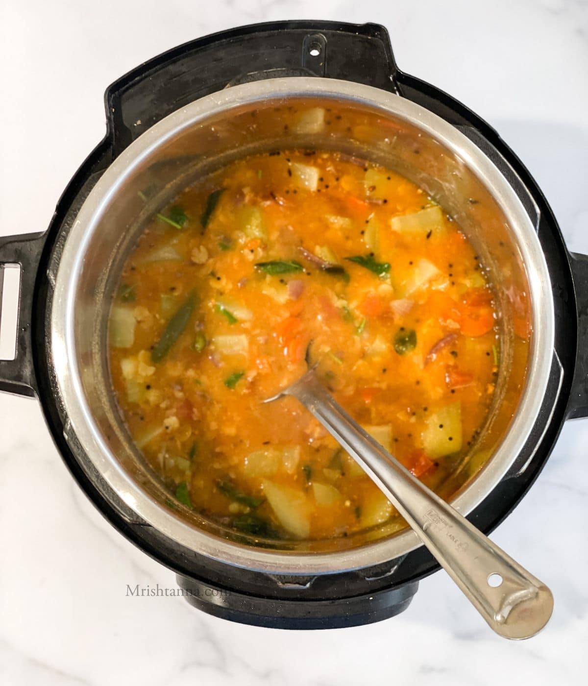 An instant pot is with vegetable sambar and a steel ladle is inserted.