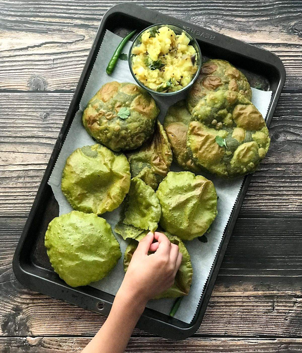 A tray is filled with fried spinach puri and a kid hand is picking up one from tray.