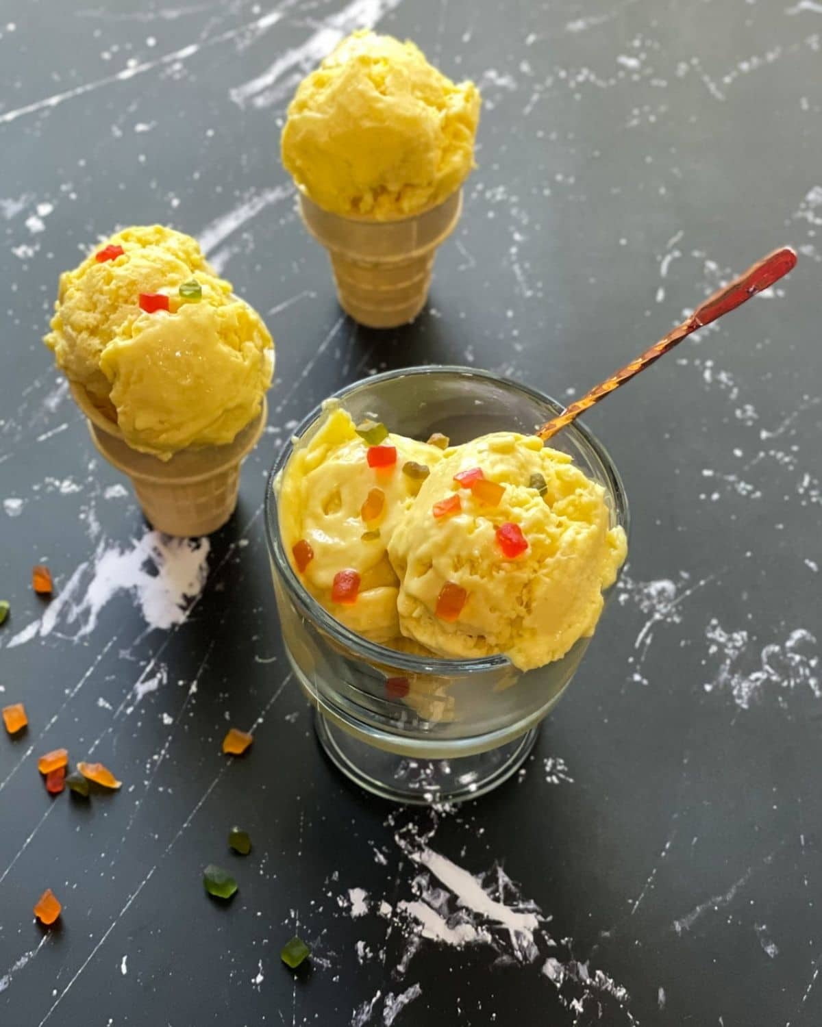 A glass bowl is with mango coconut ice cream on the table along with two ice cream cones 