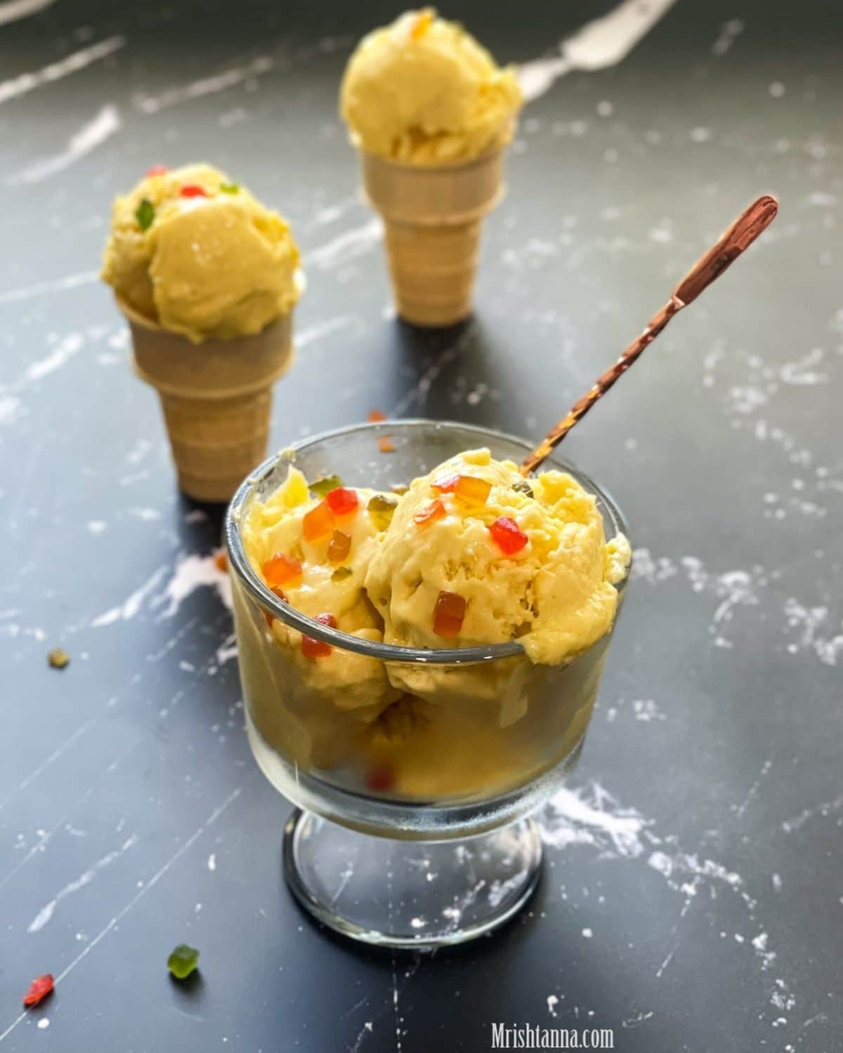 A glass bowl is with mango Ice Cream is on the table and topped with tutti fruit-ti