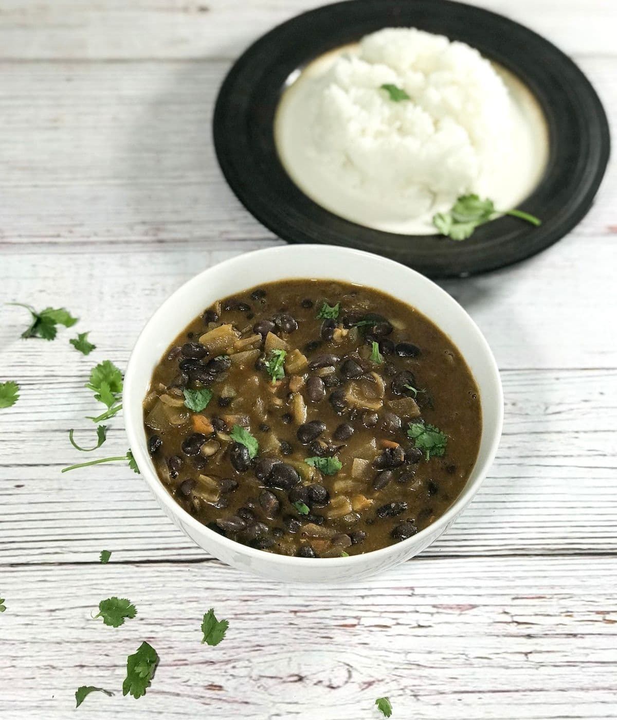 A white bowl is with black bean curry on the table along with plate of rice