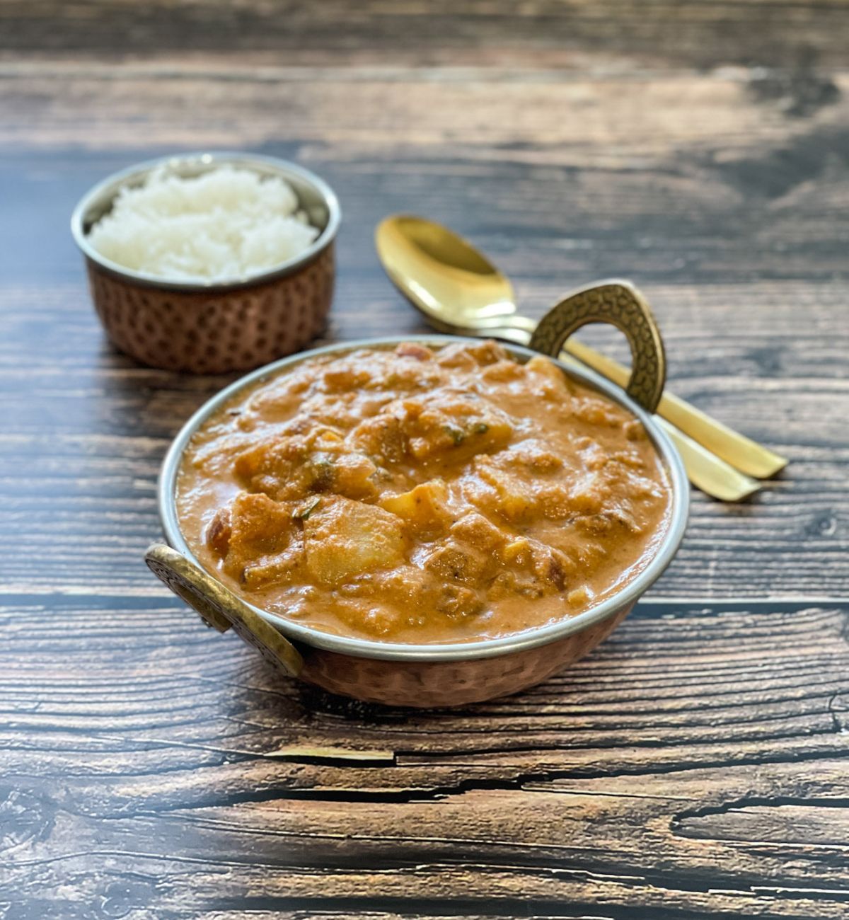 A copper bowl is with potato kurma on the table.