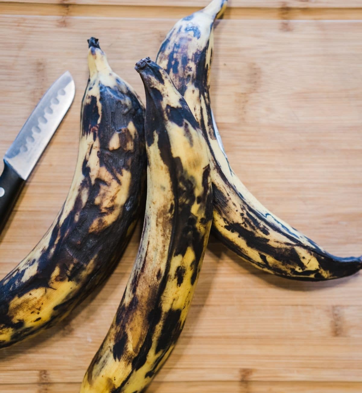 three ripe dark skinned plantains are placed on the cutting board with knife.