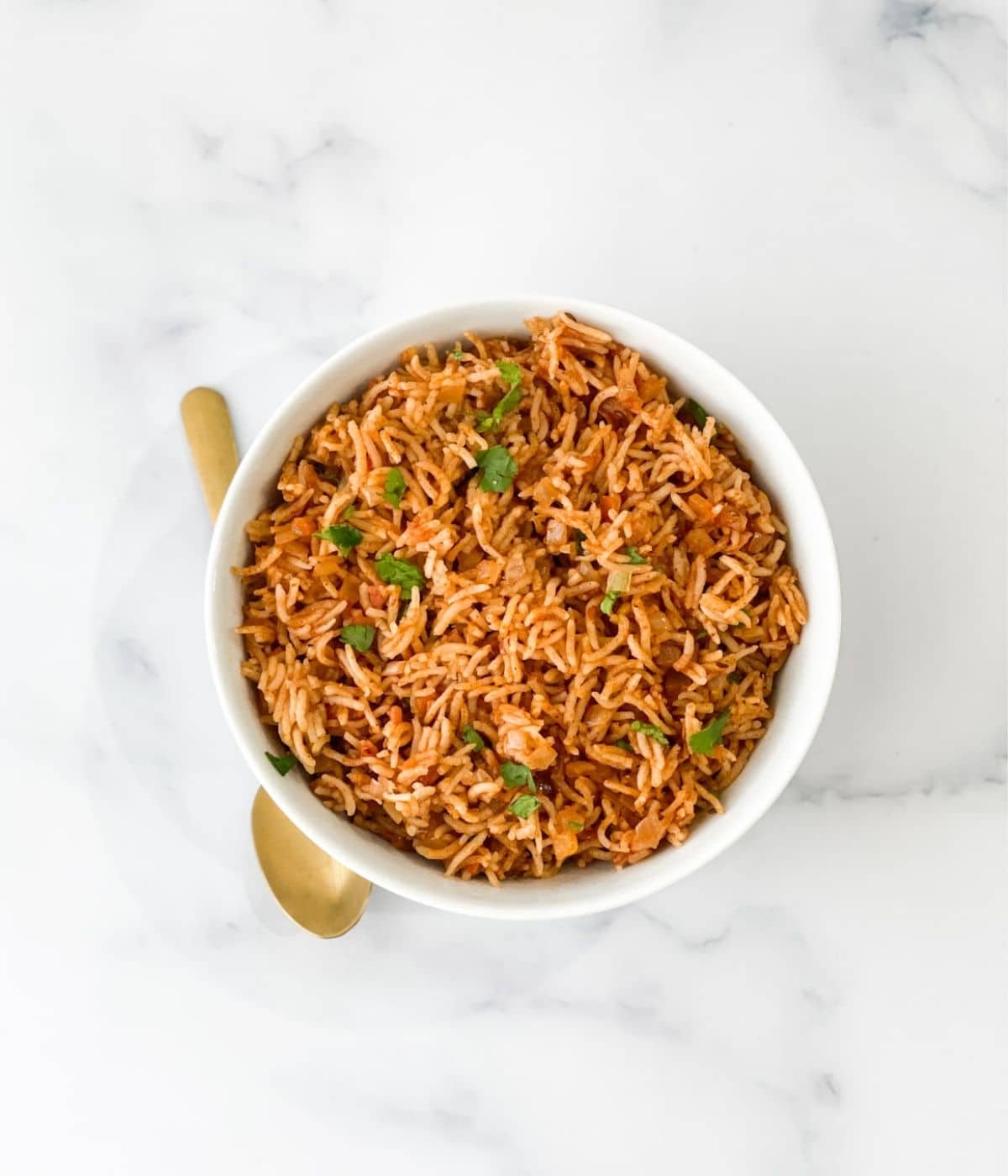 A white bowl is with tomato rice along with golden spoon by side.