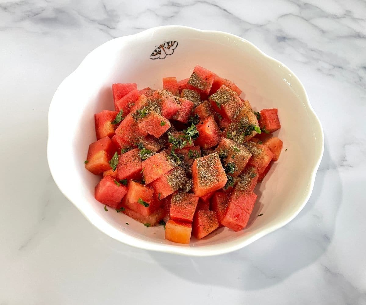 A ceramic bowl is with cubed water melon and spices for chaat.
