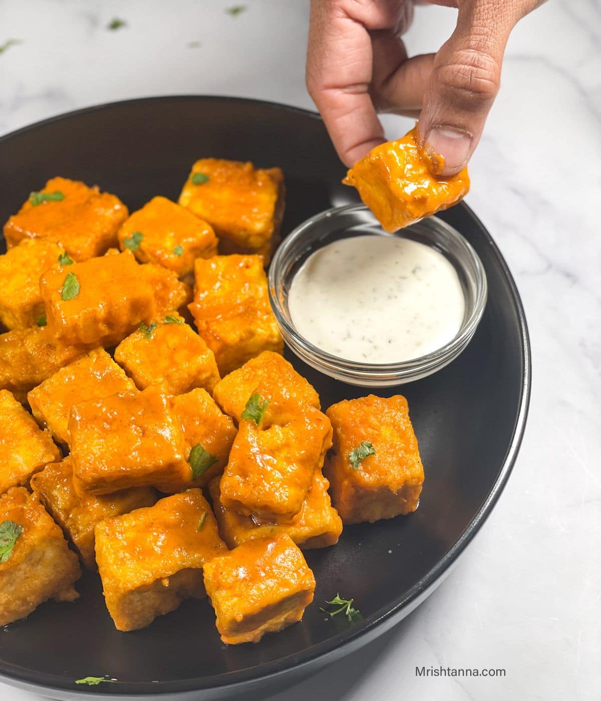 A hand is dipping the air fried buffalo tofu into sauce.