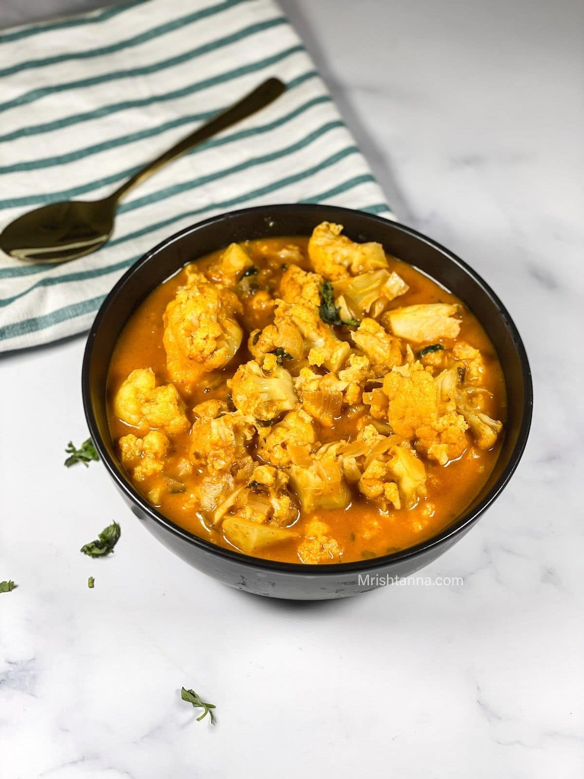 A bowl of cauliflower curry is on the table along with a golden spoon.