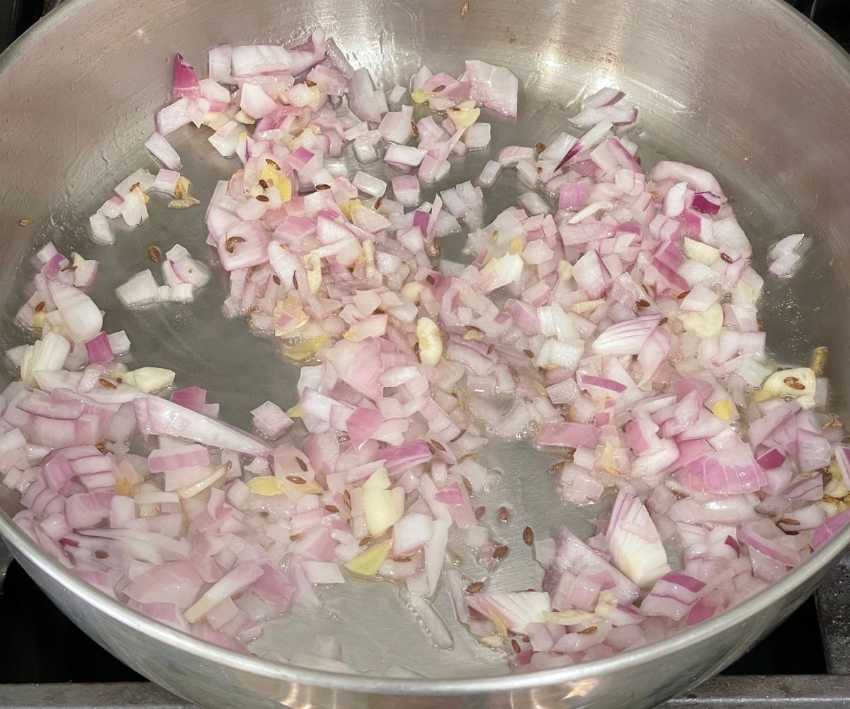 A steel pot is with cumin seeds and onions over the heat.