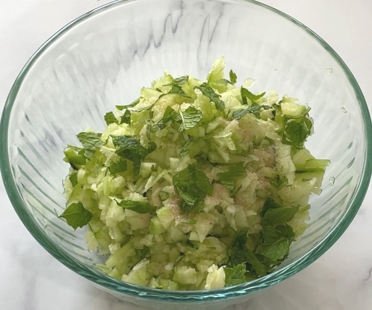 A bowl is with chopped cucumber, mint leaves on the table.