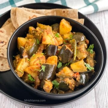 A ceramic bowl of eggplant potato curry is with chapati on the plate.