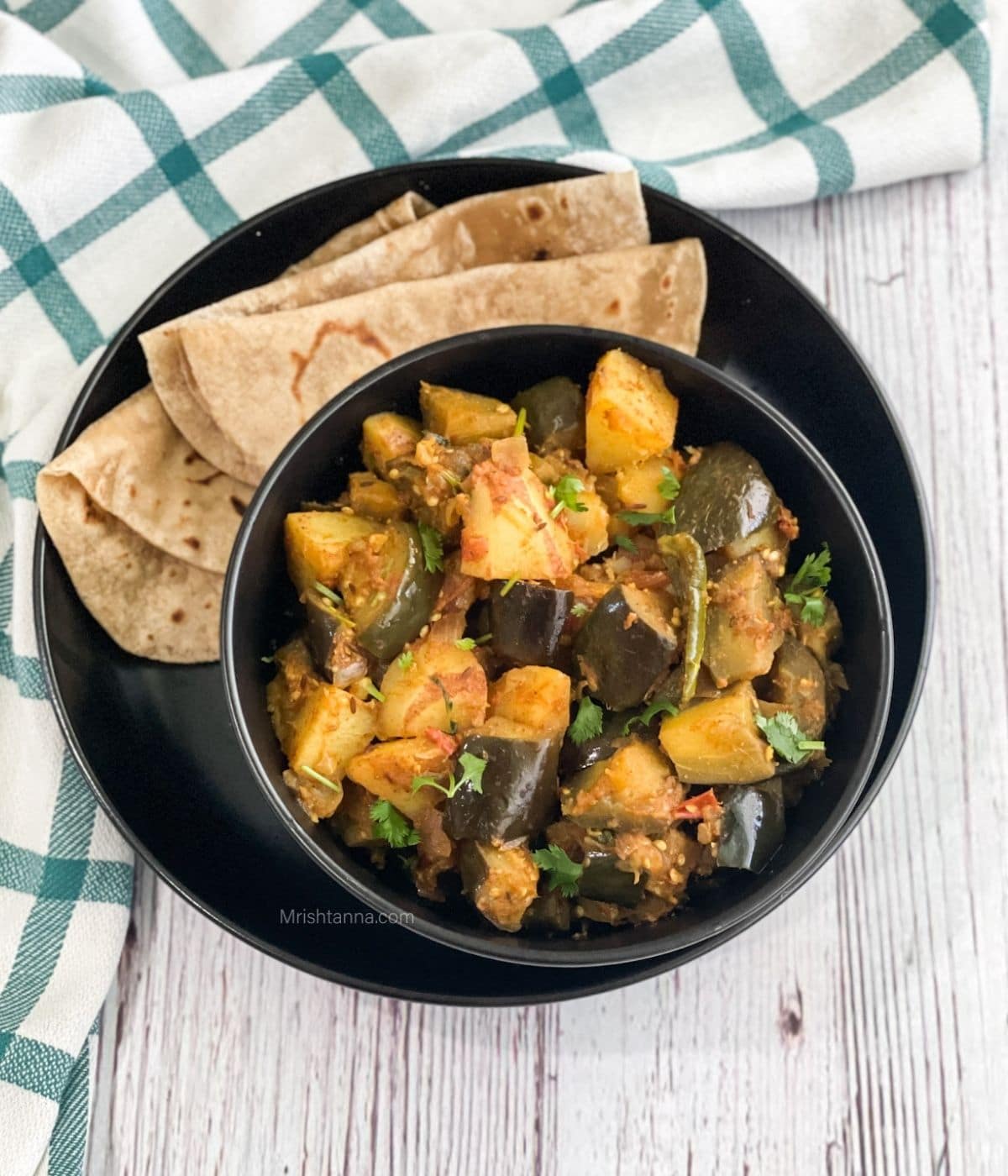 A bowl of aloo baingan sabji is on the plate with chapati.