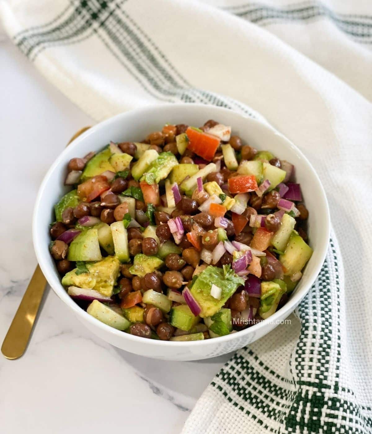 A bowl of black chana chaat is on the table with spoon on the side.