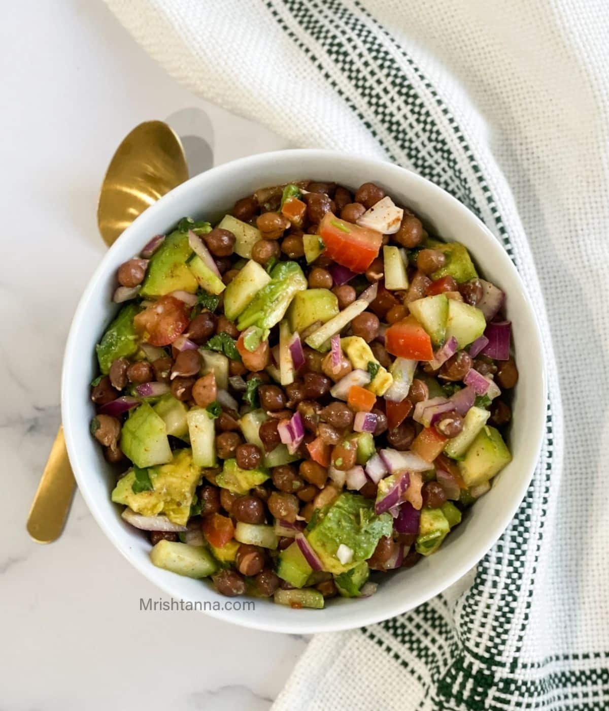 A bowl of kala chana chaat is on the marble top with napkin and spoon.