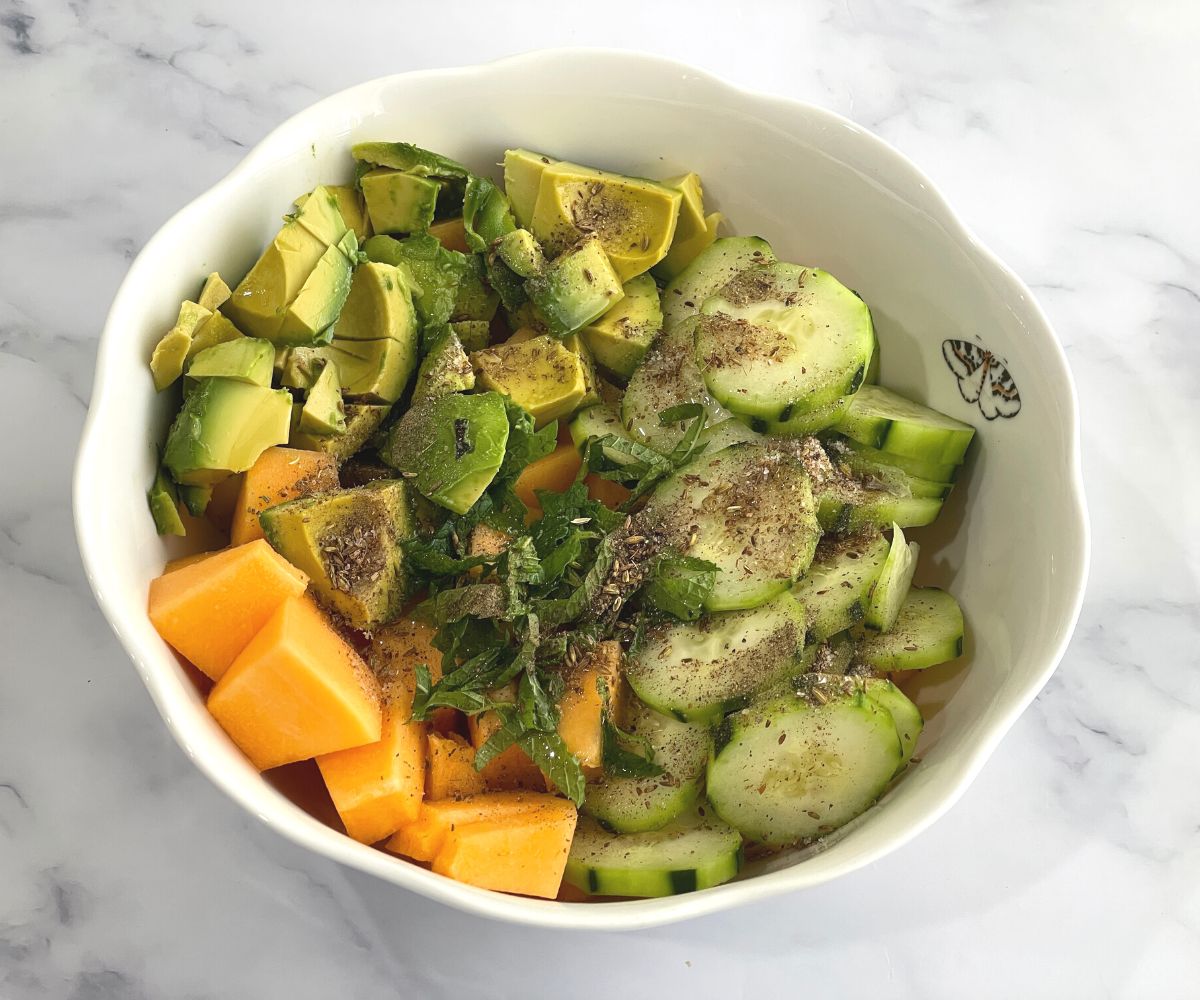 A bowl is with chopped cantaloupe, cucumber, avocados and spices for salad.