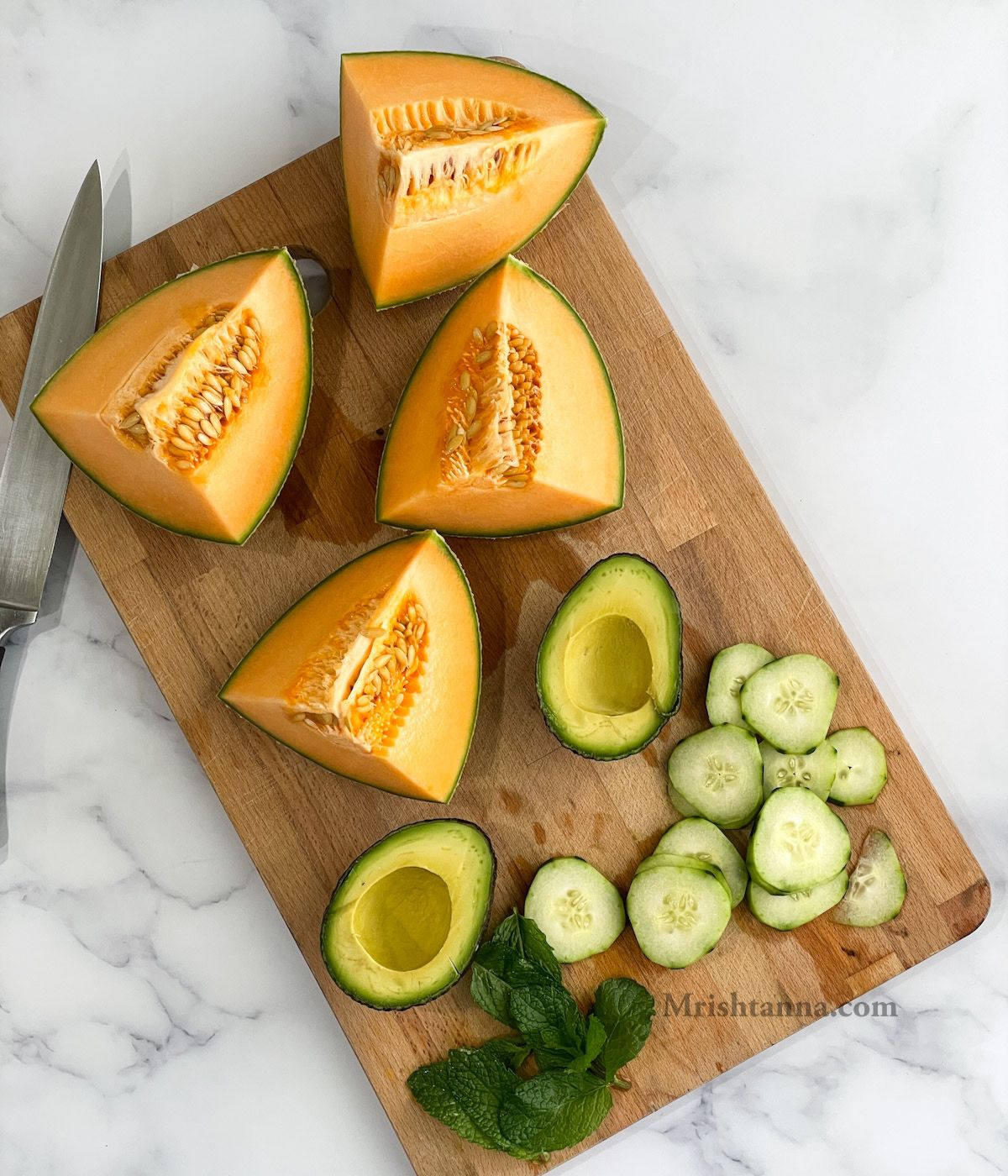 A chopping wooden board is with cantaloupe, avocados, and cucumbers.
