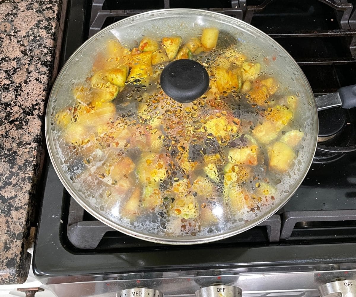 A pan of aloo baingan masala is on the heat with lid covered.